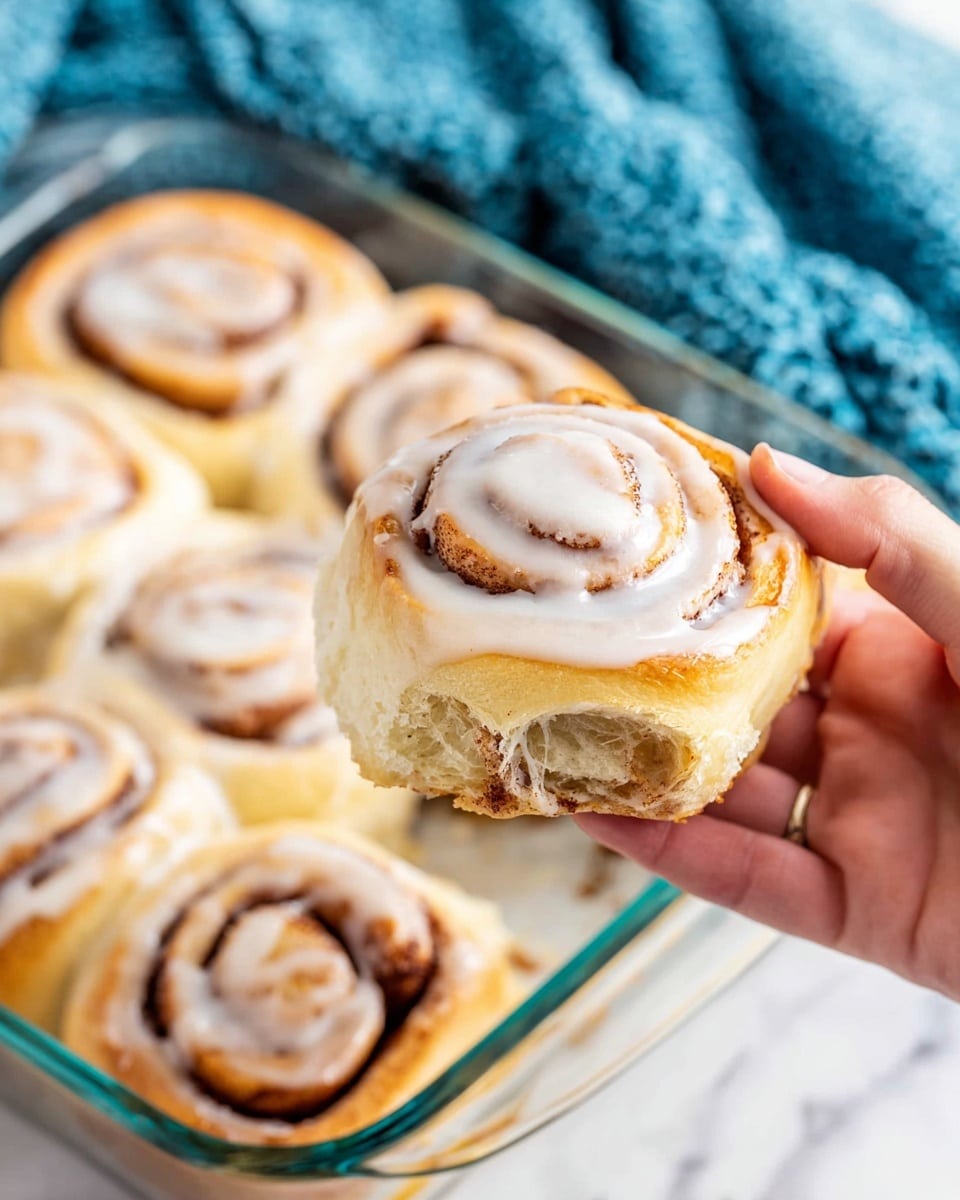 A woman's hand is holding a single cinnamon roll covered in white icing, showing its spiral shape with light brown layers of dough and cinnamon. Below, in a clear glass baking dish on a white marbled surface, there are several more cinnamon rolls also covered in icing, each with visible swirls of cinnamon and soft, golden-brown dough. A blue textured cloth is seen in the background behind the dish. photo taken with an iphone --ar 4:5 --v 7
