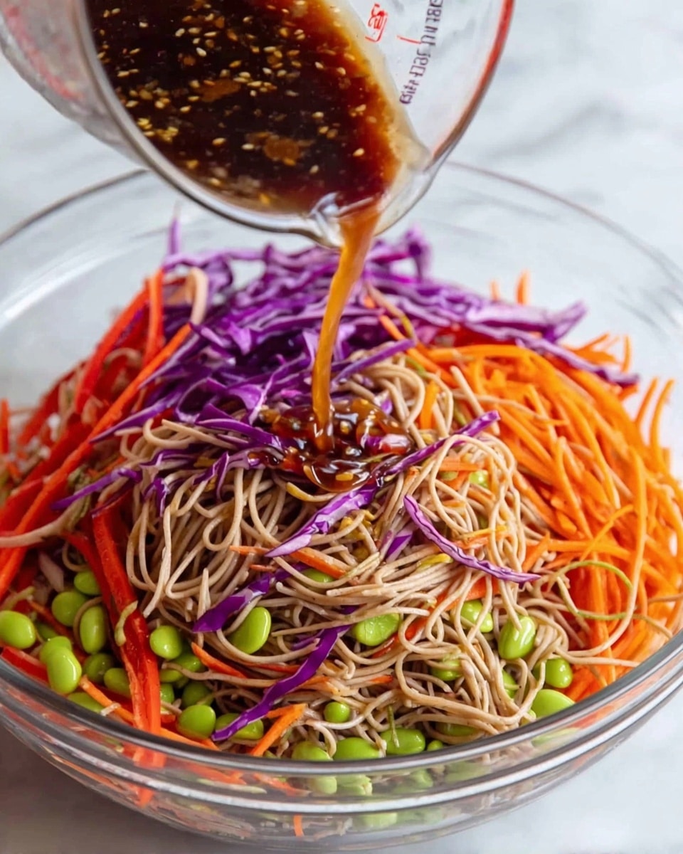 A clear glass bowl filled with three main layers of a noodle salad: the bottom layer has thin strands of light brown noodles, mixed with bright green edamame beans scattered throughout; the middle layer consists of thin strips of vibrant purple cabbage and orange shredded carrots, spread evenly over the noodles; the top layer features some red bell pepper strips for extra color. A dark brown sauce with visible small bits is being poured from a glass measuring cup over the noodles and vegetables. The bowl is placed on a surface with a white marbled texture. photo taken with an iphone --ar 4:5 --v 7