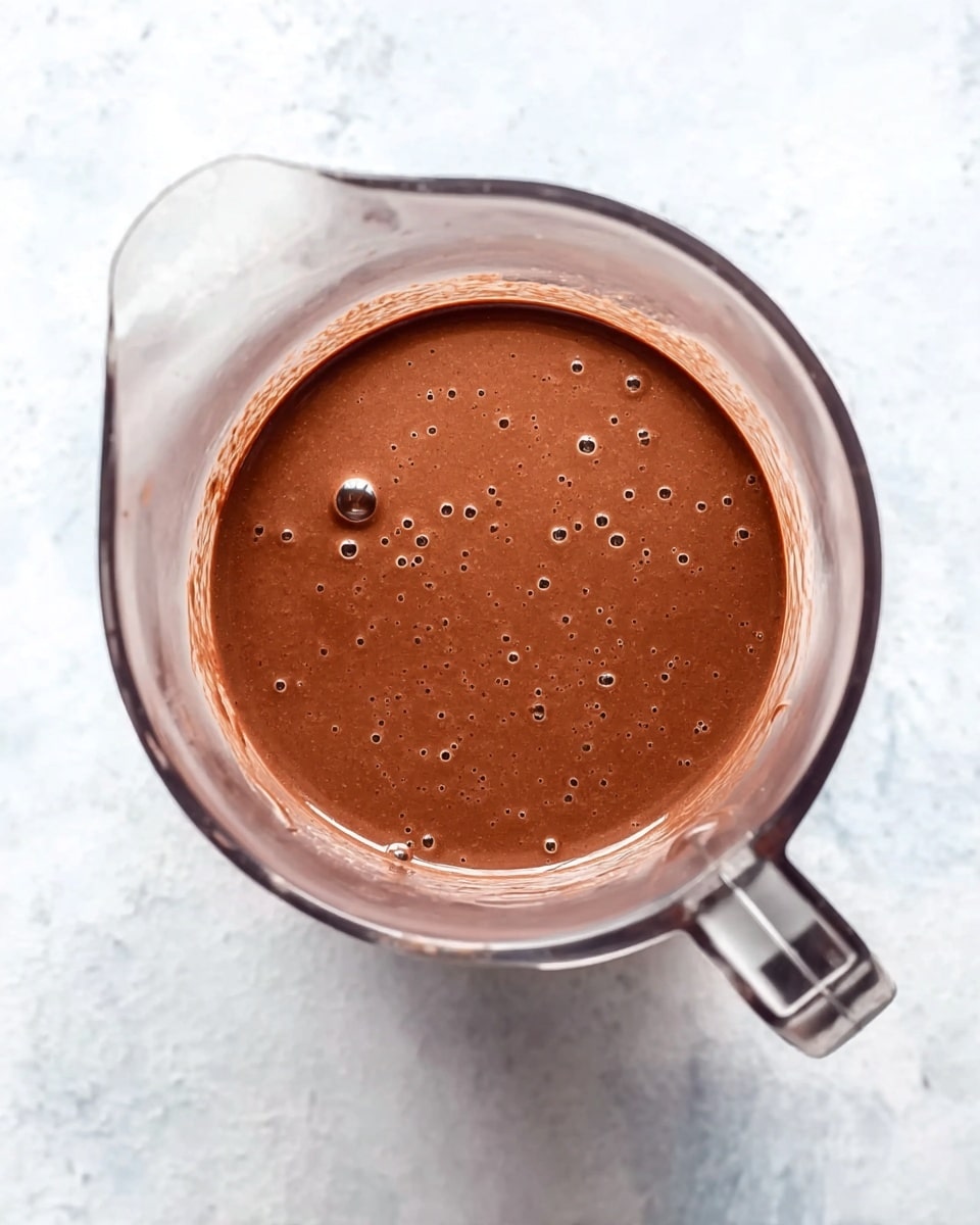 A clear blender container filled with smooth, thick chocolate batter that has a rich brown color with a few small bubbles on the surface, sitting on a white marbled texture. The blender handle is visible on the right side of the container. photo taken with an iphone --ar 4:5 --v 7