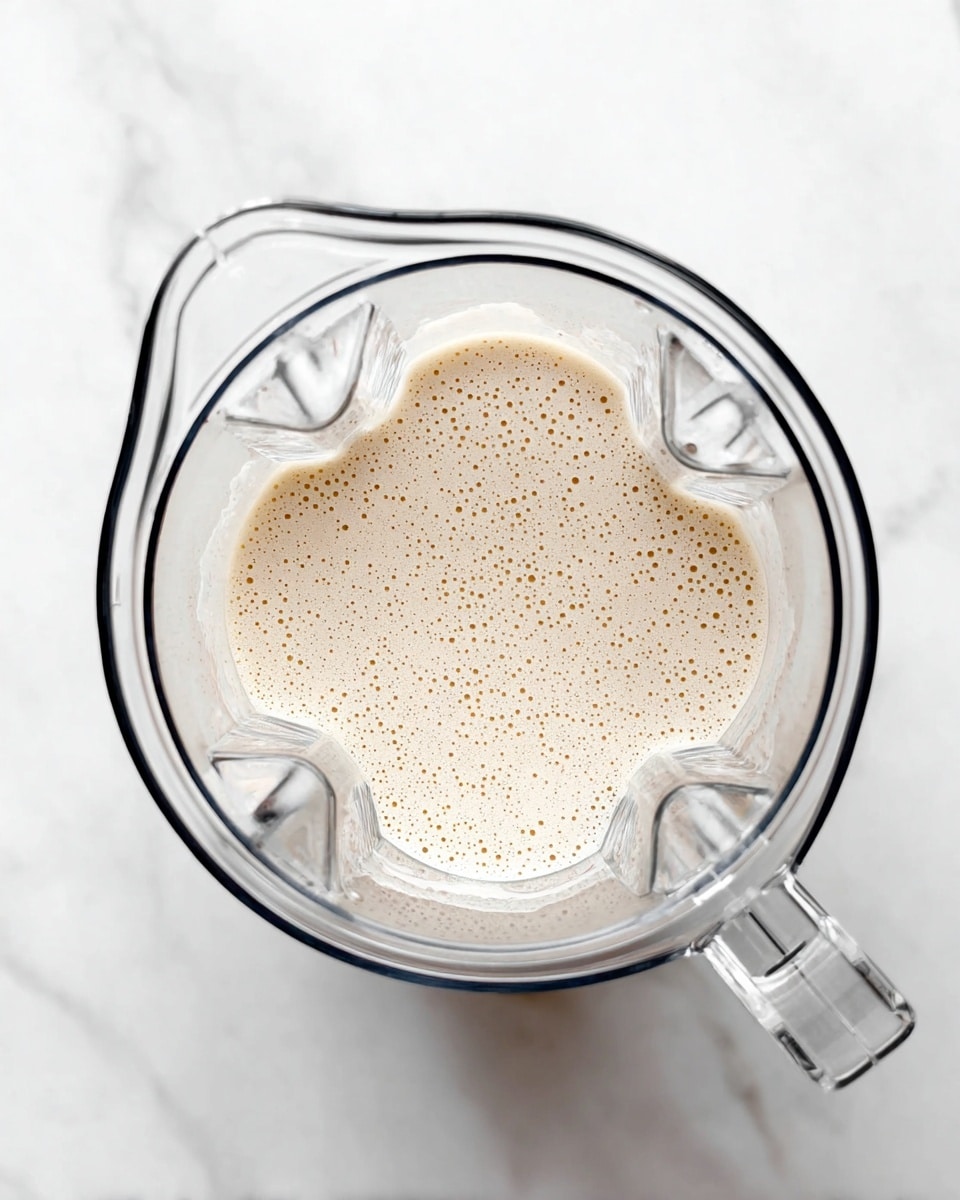 A clear blender jar viewed from the top sits on a white marbled surface, filled about halfway with a light beige liquid that has a frothy, bubbly texture on the surface. The blender jar has a handle on the right side and visible sharp blades at the bottom inside. The liquid looks smooth and creamy, with tiny bubbles spread evenly. Photo taken with an iphone --ar 4:5 --v 7