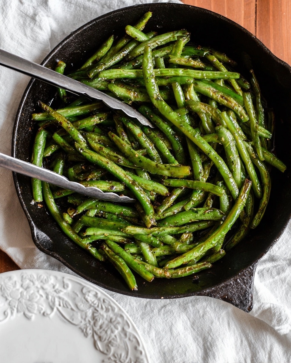 A close-up view of a black cast iron pan filled with cooked green beans, some beans showing light brown char marks and a slight glossy texture from oil and seasoning, with a pair of metal tongs resting on top; the pan sits on a wooden surface next to a crumpled white cloth and a white plate with an embossed floral pattern, all set on a white marbled texture background. photo taken with an iphone --ar 4:5 --v 7