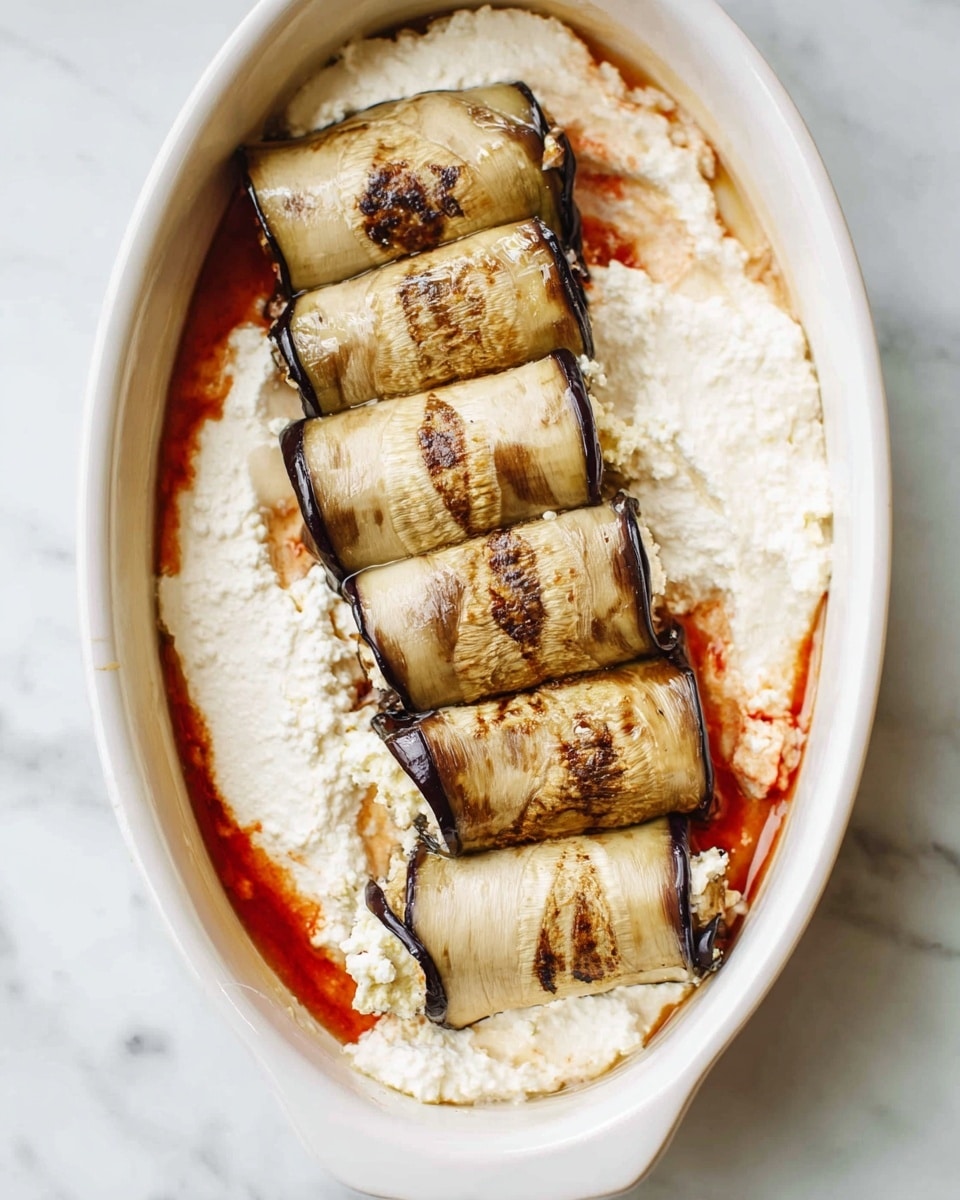 The image shows a white dish filled with a base layer of creamy, white ricotta cheese spread evenly across the bottom. On top of this base, there are four rolled eggplant slices, placed side by side in a neat row. Each eggplant slice has a light brown and beige color with dark grill marks and seeds visible, showing a soft, slightly charred texture. Around the edges of the dish, a thin reddish sauce is visible, contrasting softly with the creamy white ricotta. The dish is set on a white marbled surface. Photo taken with an iphone --ar 4:5 --v 7