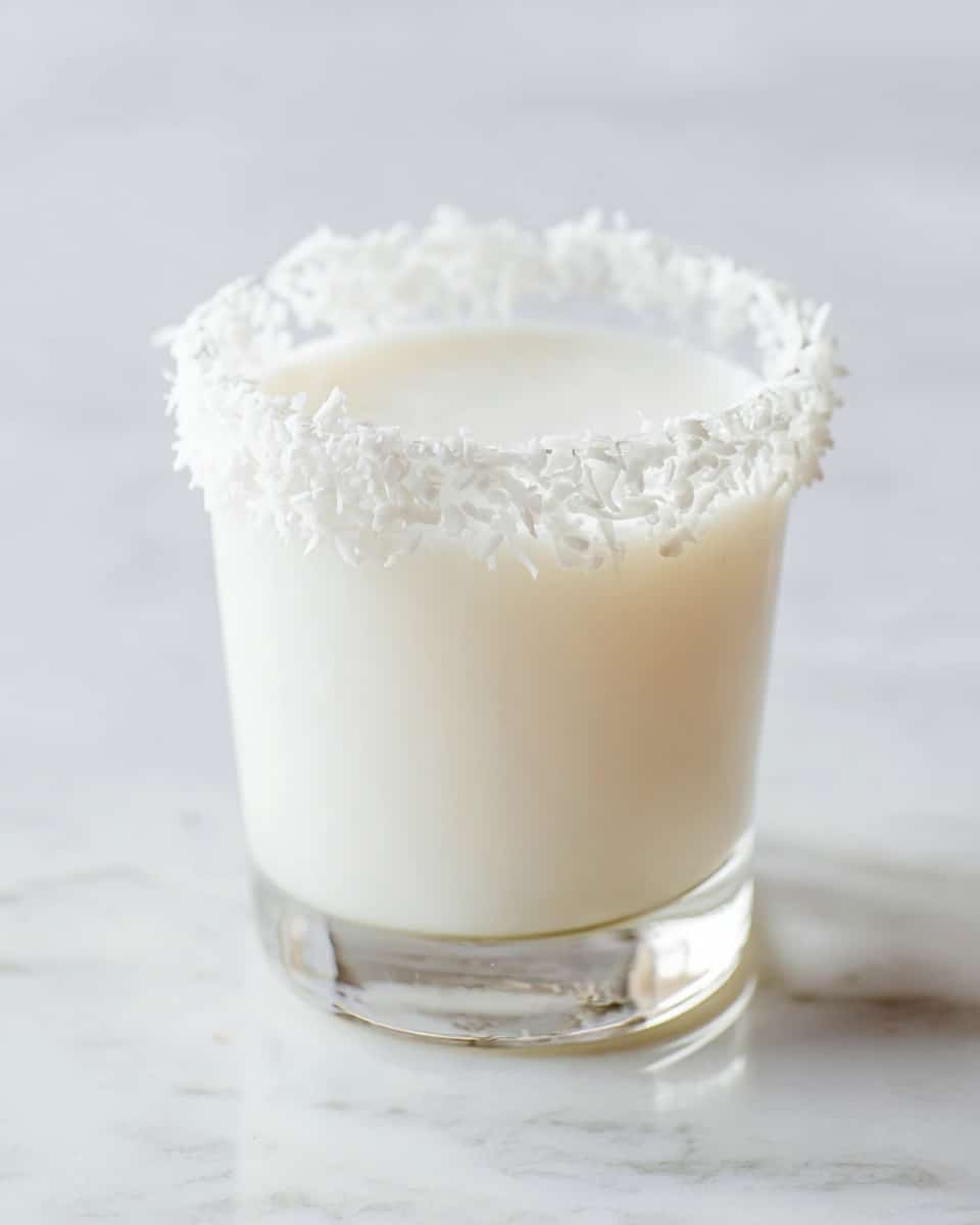 A clear glass filled close to the top with a smooth, creamy white liquid, showing a soft frothy surface; the rim of the glass is decorated with a thick layer of white shredded coconut that creates a textured and slightly fluffy border all around; the glass is placed on a white marbled surface with soft natural light highlighting the clean and fresh look of the drink photo taken with an iphone --ar 4:5 --v 7