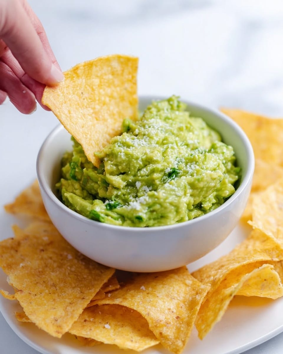 A close-up of a white bowl filled with chunky green guacamole, showing visible pieces of avocado and herbs. A woman's hand is holding a golden-yellow, slightly curved tortilla chip with coarse salt on top, dipping it into the guacamole. Around the bowl, scattered golden-yellow tortilla chips are placed on a white plate resting on a white marbled surface. The scene is bright and clean, highlighting the contrast between the creamy green guacamole and the crispy chips. Photo taken with an iphone --ar 4:5 --v 7