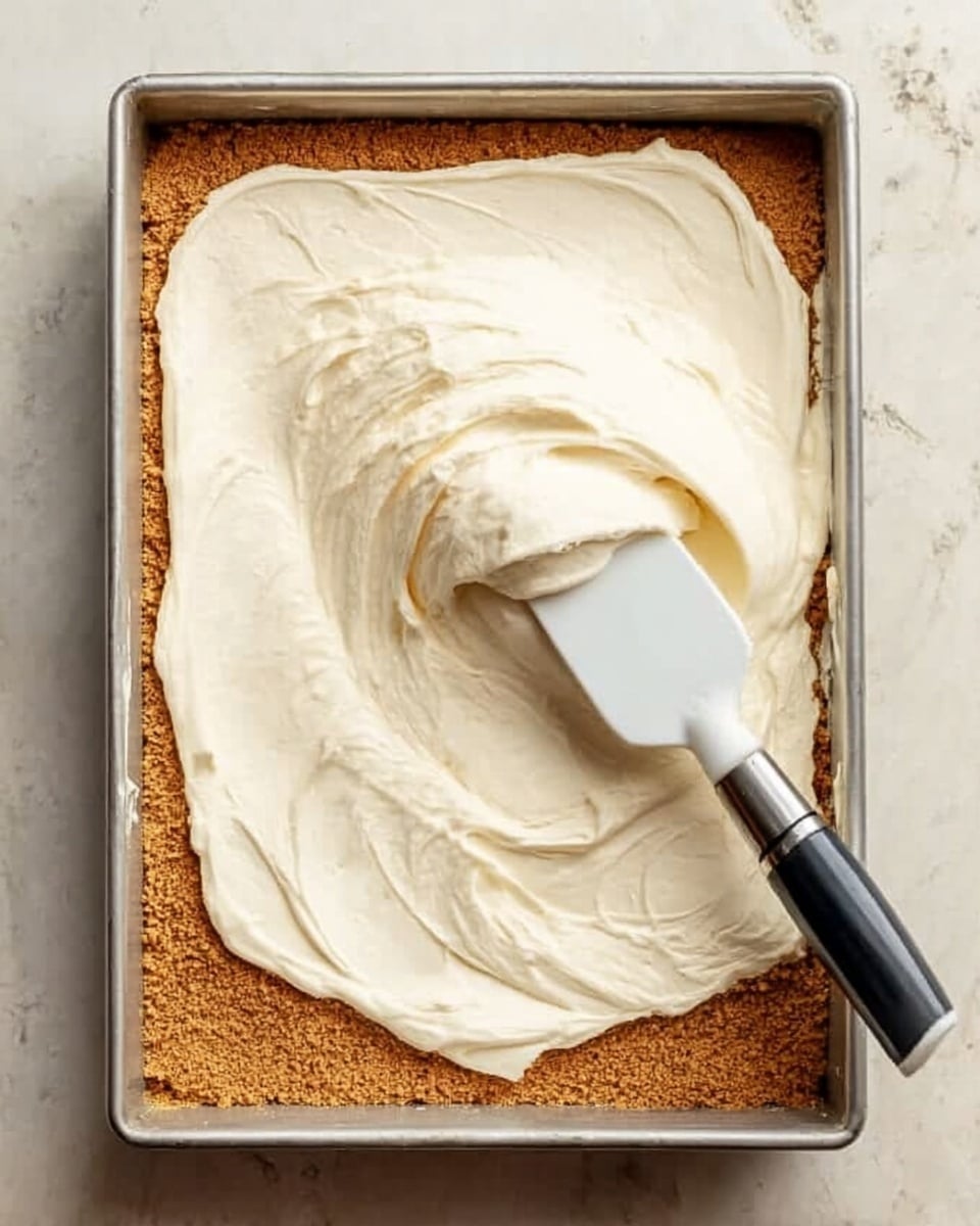 A rectangular metal dish holds a crumbly, light brown crust as the bottom layer, with a thick, creamy white layer being spread on top using a white spatula with a silver handle. The spatula is placed near the right edge of the dish, smoothing the creamy layer in soft swirls. The dish is set on a flat surface with a white marbled texture. Photo taken with an iphone --ar 4:5 --v 7