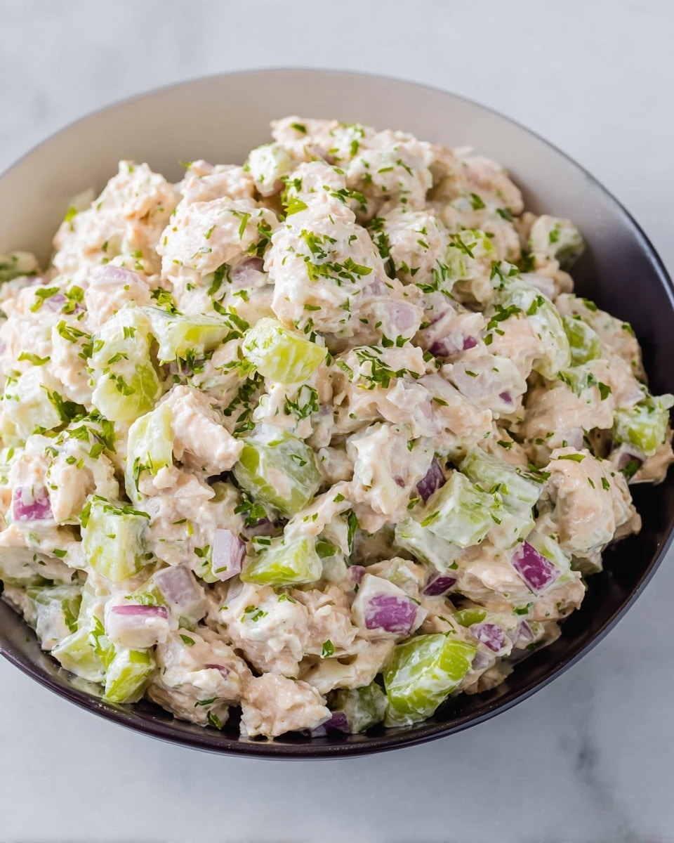 A close-up view of a chicken salad in a white bowl on a white marbled surface, showing three main layers: the base layer of light creamy chicken pieces mixed with chopped green celery pieces giving a firm texture, the middle layer of diced pale purple onions mixed evenly throughout the salad adding color contrast, and the top layer sprinkled with finely chopped green herbs lending freshness; the salad looks creamy with bits of white dressing visible coating the chunky ingredients photo taken with an iphone --ar 4:5 --v 7