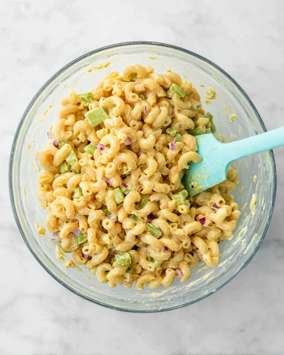 A clear glass bowl filled with creamy yellow macaroni salad mixed evenly with small green celery pieces and bits of red onion, giving a fresh, crunchy look. The macaroni is elbow-shaped with a smooth, glossy texture from the dressing. A light blue spatula is partially inside the bowl, resting on the right side, with the salad clinging to it. The bowl sits on a white marbled surface, bright and clean. photo taken with an iphone --ar 4:5 --v 7
