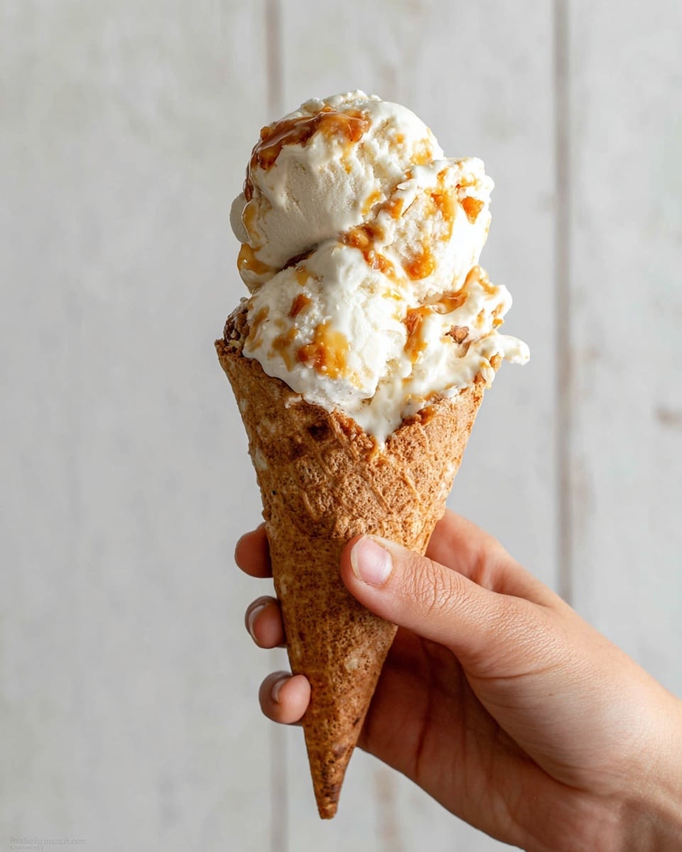 A woman's hand holds a waffle cone with three scoops of creamy white ice cream that has swirls and patches of golden caramel and light brown bits mixed in. The ice cream looks soft and slightly melting at the edges. The cone is textured with a classic waffle pattern and has a light brown color. The background is a simple off-white wall with subtle vertical lines, and the surface beneath is a white marbled texture photo taken with an iphone --ar 4:5 --v 7