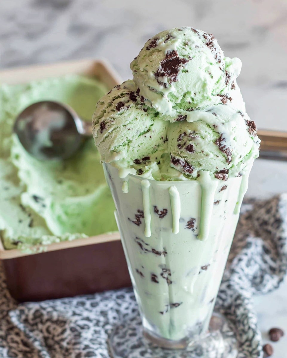 The image shows a rectangular metal container filled with bright green mint chocolate chip ice cream, which has a creamy and slightly fluffy texture. There are visible small dark chocolate pieces scattered throughout the ice cream, giving it a speckled look. The ice cream is scooped in parts, showing uneven layers and soft peaks. A closed metal ice cream scooper rests inside the container. The container is placed on a white marbled surface. photo taken with an iphone --ar 4:5 --v 7