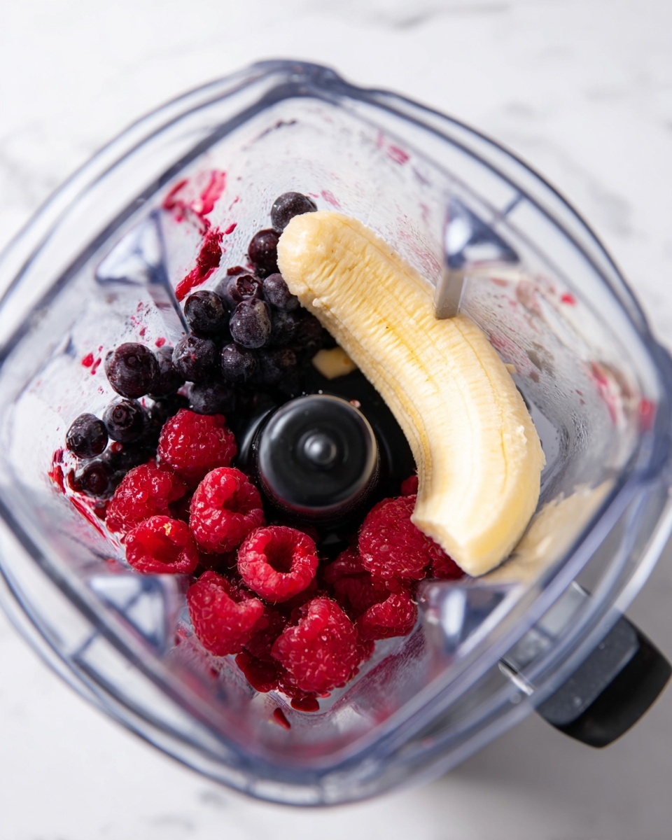The image shows a clear blender container filled with three layers of fruit: the bottom layer has small dark purple blueberries, the middle layer consists of bright red raspberries, and the top layer includes a large, partially peeled yellow banana. The black blender blade is centered among the fruits. The blender sits on a surface with a white marbled texture. Photo taken with an iphone --ar 4:5 --v 7