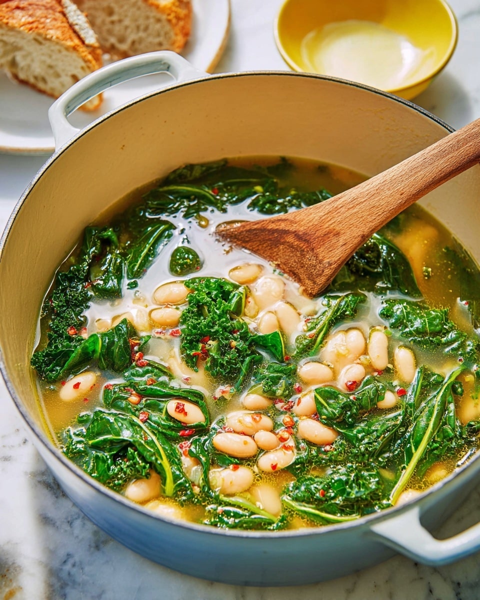A light beige pot filled with a soup showing three main layers: at the bottom, a thin broth with a pale yellow color; in the middle, many white beans scattered evenly; on top, green kale leaves floating and partially submerged, with visible texture and veins, sprinkled with small red chili flakes; a smooth wooden spoon rests inside the pot. The pot sits on a white marbled surface, with a white plate holding torn bread on the left and a small yellow bowl with a creamy substance behind the pot. Photo taken with an iphone --ar 4:5 --v 7