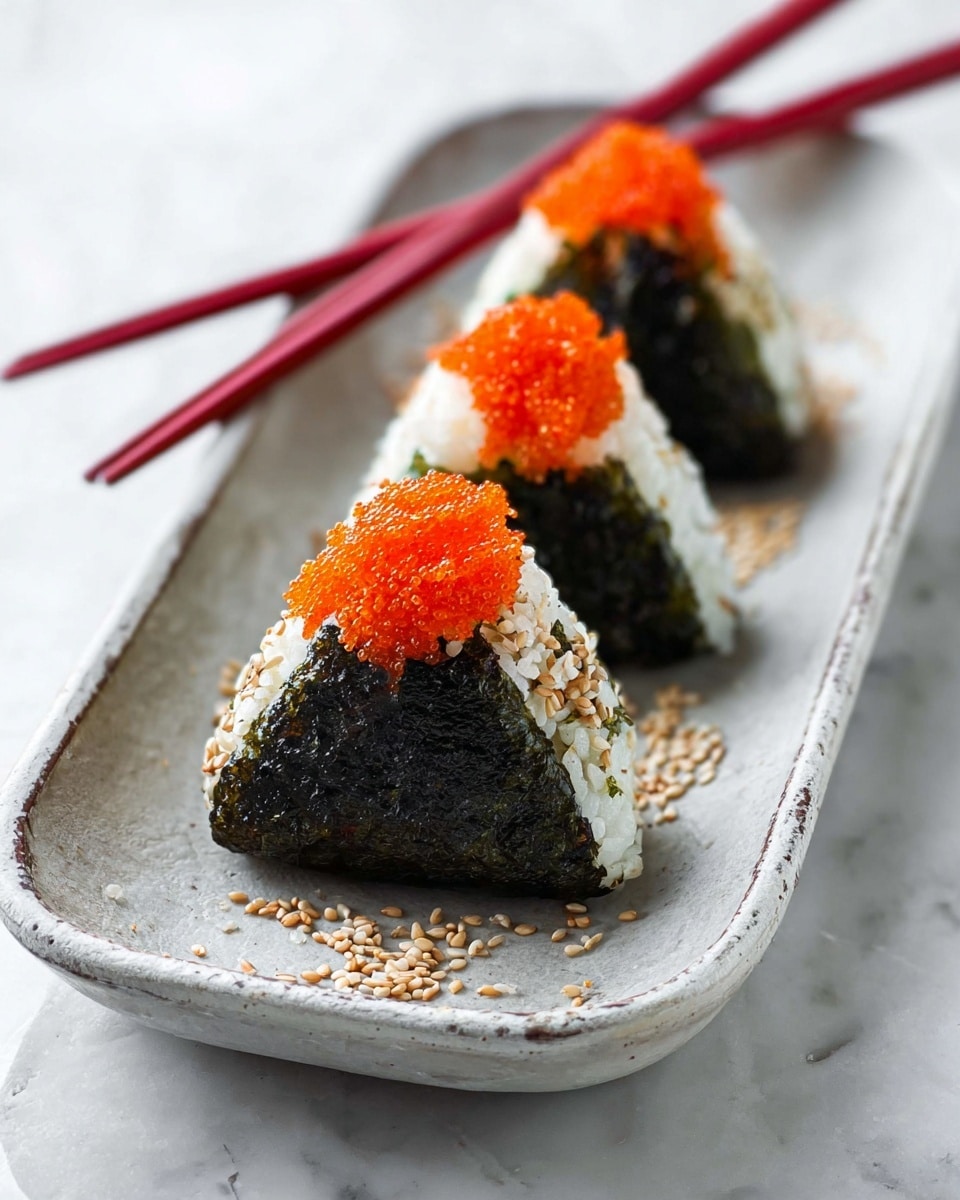 Three triangle-shaped rice balls are placed in a row on a long white plate with a slightly rough texture. Each rice ball has a bottom layer wrapped in dark green seaweed, a middle layer of white rice mixed with sesame seeds, and an orange fish roe topping on the top. Lightly scattered sesame seeds are seen around the rice balls on the plate. A pair of red chopsticks rests across the top left edge of the plate. The background surface is white with a soft marbled pattern. Photo taken with an iphone --ar 4:5 --v 7