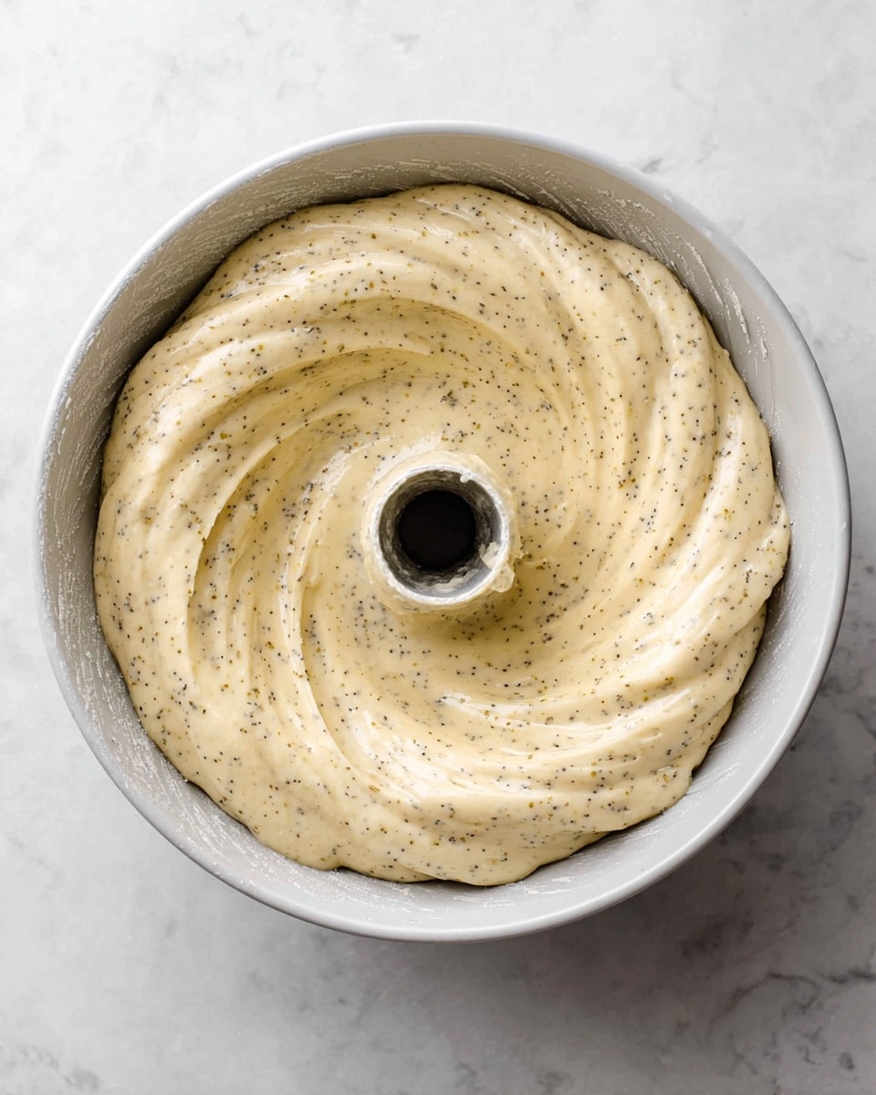 A white Bundt pan filled with pale yellow cake batter mixed with small black specks is shown from above. The batter is evenly spread, filling the pan almost to the top, with gentle swirls and smooth texture visible. The white marbled surface is seen under the pan. Photo taken with an iphone --ar 4:5 --v 7
