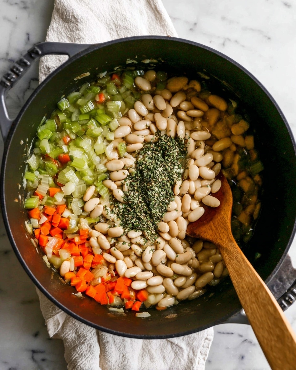 A black cooking pot with three visible layers inside shows a mix of soft cooked chopped vegetables including green celery and orange carrot evenly spread on one side, a cluster of light beige white beans in the center with dried green herbs sprinkled on top, and a wooden spatula resting on the right side inside the pot, all set on a white marbled surface with a white cloth towel beneath the pot photo taken with an iphone --ar 4:5 --v 7