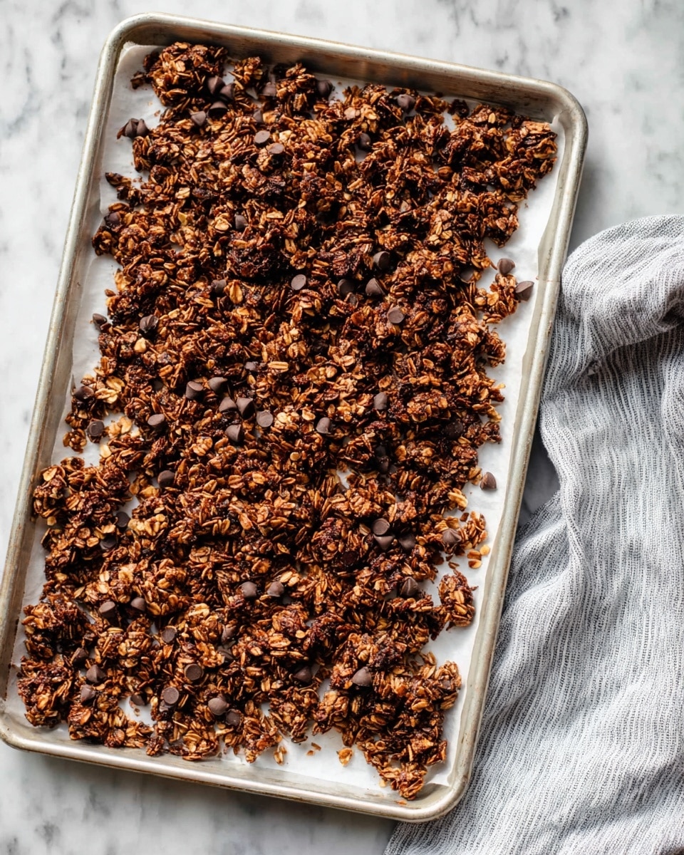 A baking tray with a white sheet of parchment paper holds uneven clusters of dark brown granola made of oats mixed with small dark chocolate chips. The granola pieces have a rough, crunchy texture with visible oat flakes and chocolate chips scattered throughout. The tray sits on a white marbled surface, and a soft gray and white striped cloth is visible on the right side. photo taken with an iphone --ar 4:5 --v 7