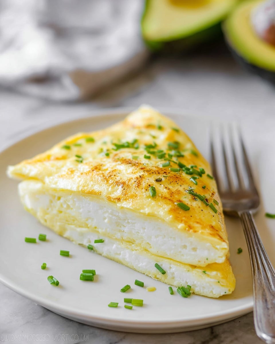 A white plate holds a folded omelette with a soft, fluffy texture that shows layers of white egg whites inside and a slightly golden-yellow egg layer on the outside. Small chopped green chives are sprinkled on top of the omelette. On the right side of the plate, there are chunks of green avocado with a creamy, smooth texture. The plate sits on a beige cloth over a white marbled surface. A silver fork is placed near the bottom left corner of the image. photo taken with an iphone --ar 4:5 --v 7