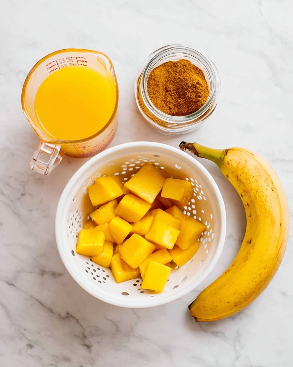 A white colander filled with bright yellow mango chunks sits in the center on a white marbled surface, next to a clear glass measuring cup filled with orange-yellow juice on the left, a small clear glass jar with brown cinnamon powder in the center-right, and a whole yellow banana with a green tip on the far right, all items neatly arranged in a row photo taken with an iphone --ar 4:5 --v 7