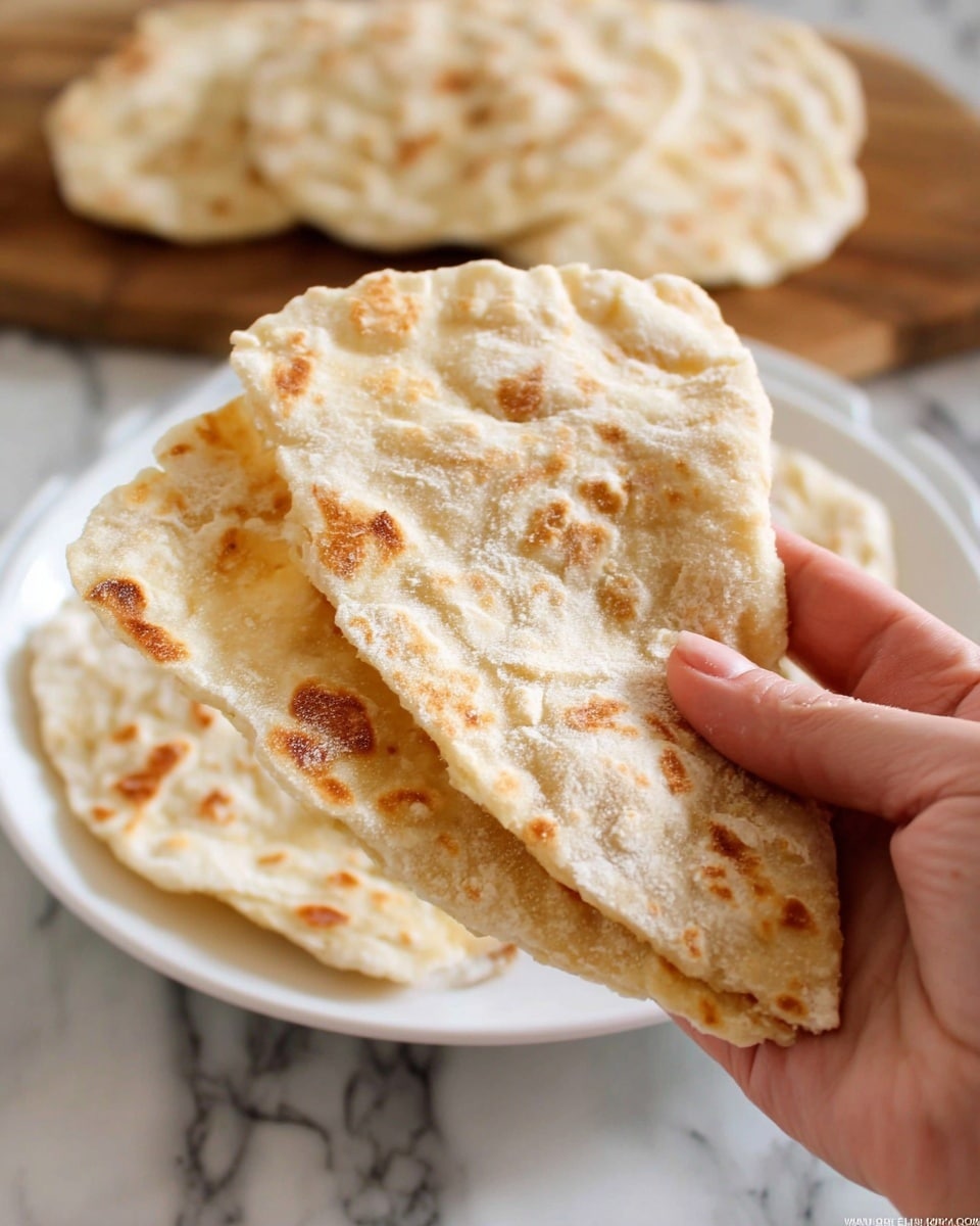 A woman's hand holds two folded flatbreads with light golden brown spots and a soft, slightly powdery texture. In the background, more flatbreads lay on a white plate on a white marbled surface. The flatbreads are thin and have a slightly uneven, homemade look with some puffiness and darker toasted areas. Photo taken with an iphone --ar 4:5 --v 7