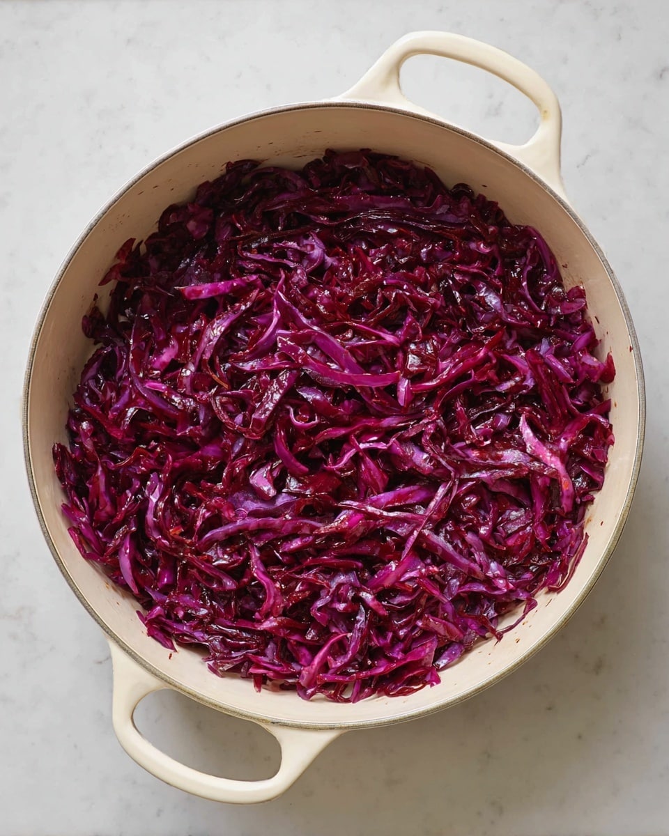 A large white pot with two handles holds a single thick layer of cooked red cabbage. The cabbage is finely shredded and has a deep purple-red color with a shiny, soft texture, spread evenly inside the pot. The pot rests on a white marbled surface. photo taken with an iphone --ar 4:5 --v 7