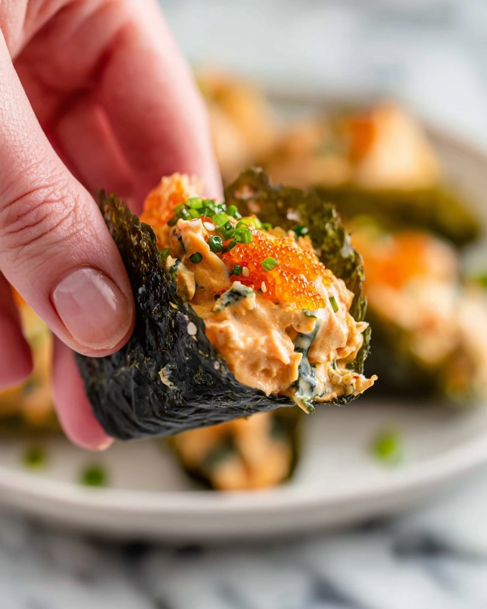 A close-up of a woman's hand holding a small sushi taco made with dark green seaweed as the base layer, filled with a creamy light orange mixture of seafood and mayonnaise, topped with bright orange fish eggs and sprinkled with small green herb bits and white sesame seeds, showing a mix of soft and slightly textured layers. The background features a white marbled surface with a blurred white plate holding more of the sushi tacos. Photo taken with an iphone --ar 4:5 --v 7