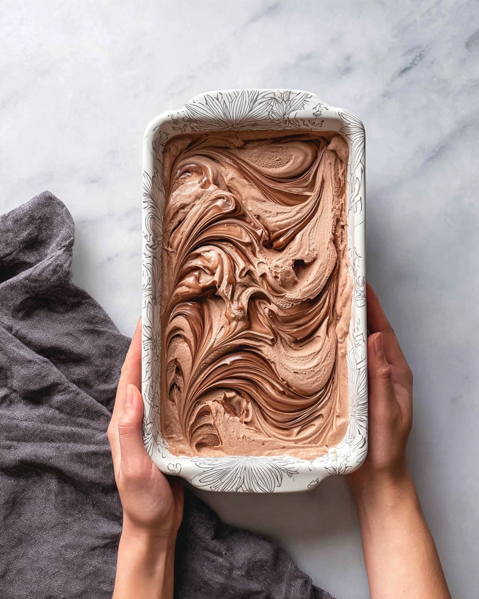 A rectangular white dish with gray floral patterns is filled with swirled chocolate ice cream showing two layers: a light brown creamy chocolate layer mixed with darker rich chocolate swirls on top, creating a smooth, flowing marbled texture. The dish is held by a woman's hand at the top and another woman's hand at the bottom, resting against a white marbled surface with a folded dark gray cloth nearby. The overall look is inviting and smooth with a mix of soft and glossy finishes photo taken with an iphone --ar 4:5 --v 7