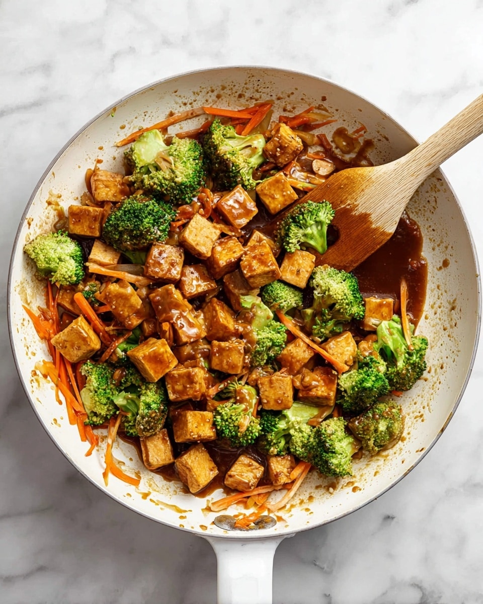 A white pan holds a cooked mix of three main layers: small golden brown cubes of tofu coated in sauce at the bottom, bright green broccoli florets scattered evenly throughout, and thin orange carrot strips spread on top, all glistening with a thick, shiny brown sauce. A wooden spatula rests inside the pan, touching the tofu and vegetables, with sauce drops around the pan edges. The pan is set on a white marbled surface. Photo taken with an iphone --ar 4:5 --v 7