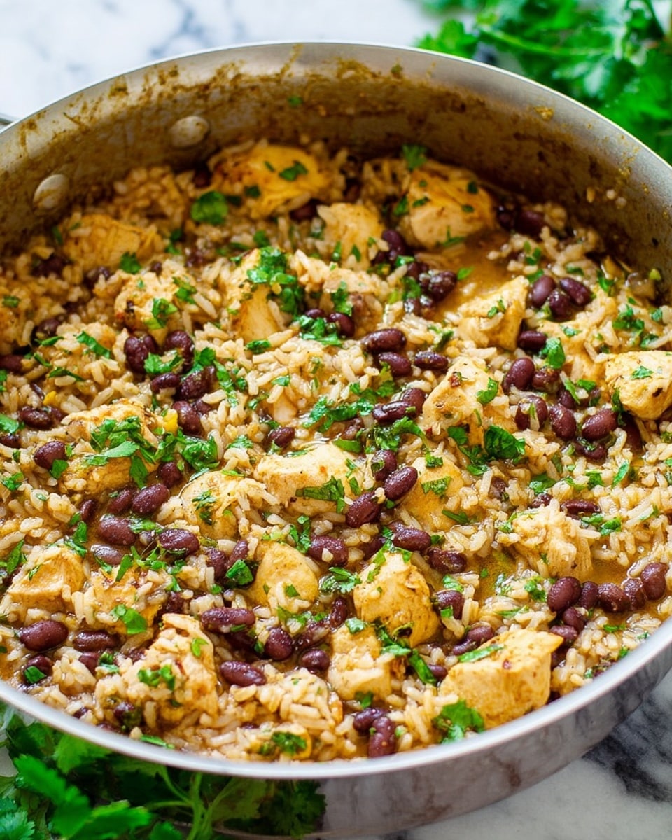 This image shows a close-up of a silver pan filled with a dish made of light brown cooked rice mixed with chunks of light yellow cooked chicken, small black beans scattered throughout, and finely chopped green herbs sprinkled on top. The food has a moist and slightly shiny texture, with the chicken pieces slightly raised above the rice. Around the pan, fresh green herb leaves are visible on a white marbled surface. The photo captures the warm colors and soft textures clearly, with the pan slightly stained on the inside edges from cooking. photo taken with an iphone --ar 4:5 --v 7