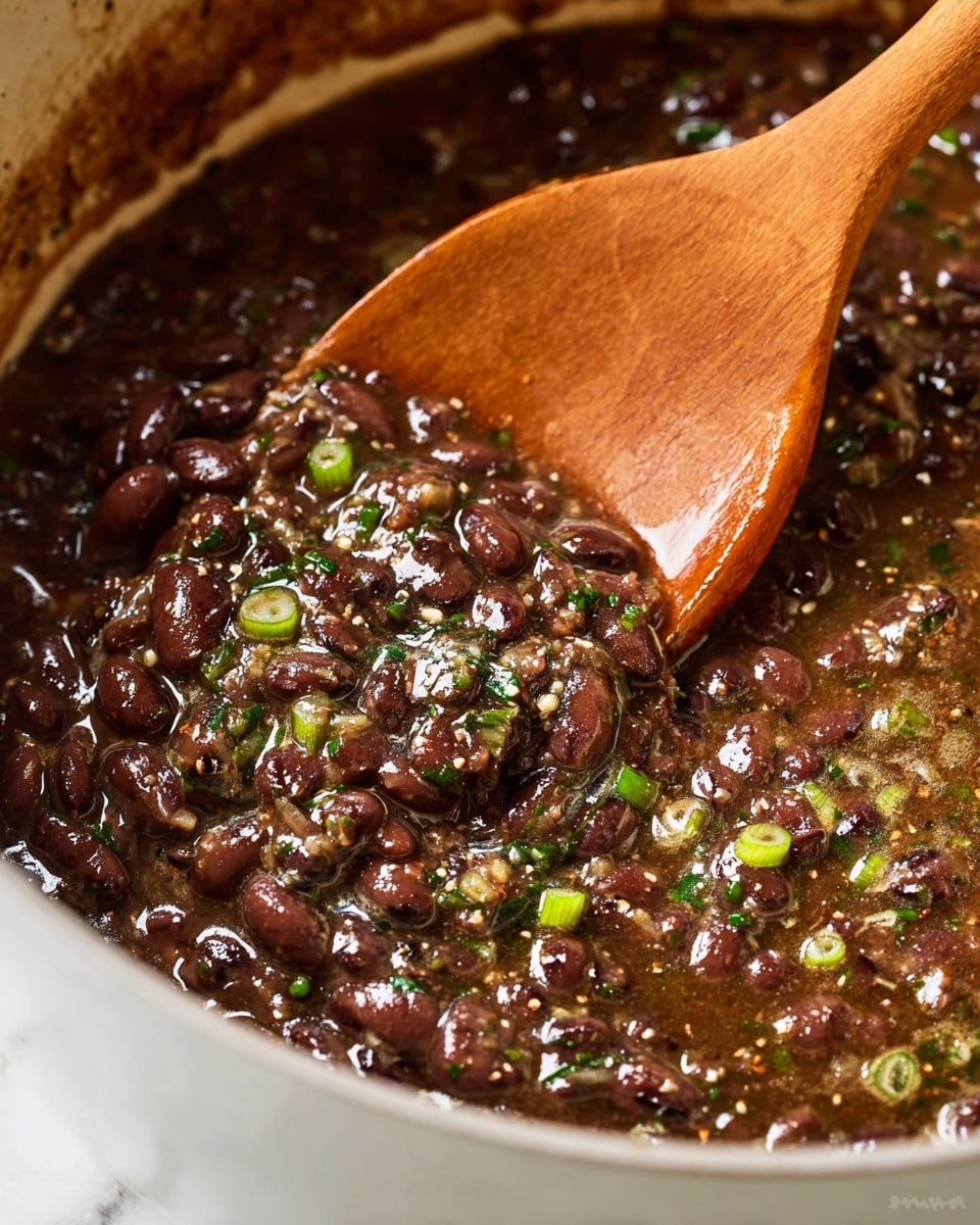 A close-up top view of a white pot filled with a thick, dark brown stew of cooked black beans with small bits of vegetables mixed in. The stew has a slightly shiny surface with scattered green herbs and thin round slices of light green onions floating on top. The pot is placed on a white marbled surface with a lemon wedge and a small bowl partially visible on the side. The edges of the pot show slight browning from cooking. Photo taken with an iphone --ar 4:5 --v 7