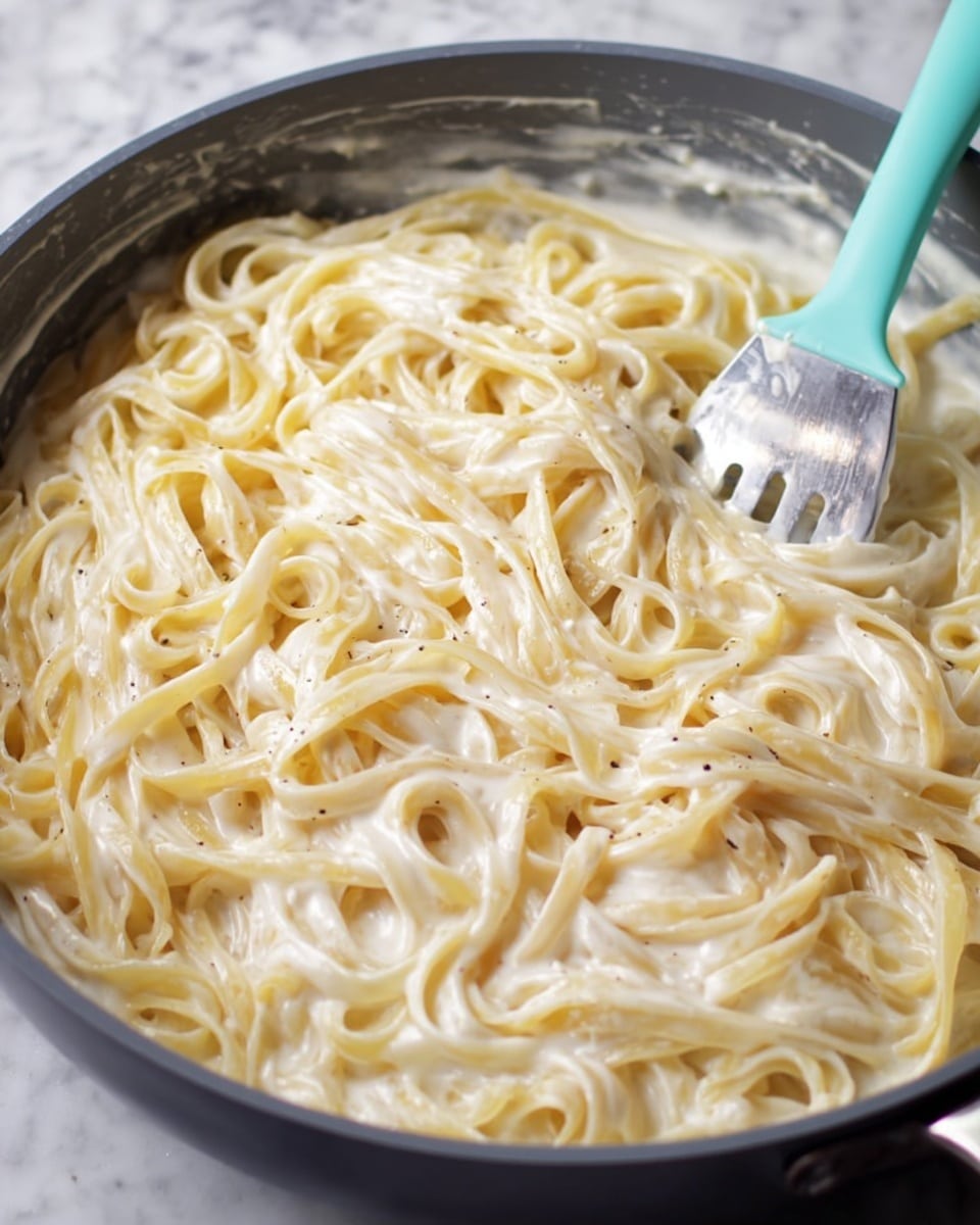 A close-up view of a pan filled with fettuccine noodles covered in a creamy white sauce, the pasta strands are soft and slightly glossy, evenly coated with the sauce that has a smooth texture with small bubbles showing slight movement. The pan is dark gray, and a mint green spoon with a metal handle is resting on the right side, partly buried in the pasta. The background is a white marbled surface. photo taken with an iphone --ar 4:5 --v 7