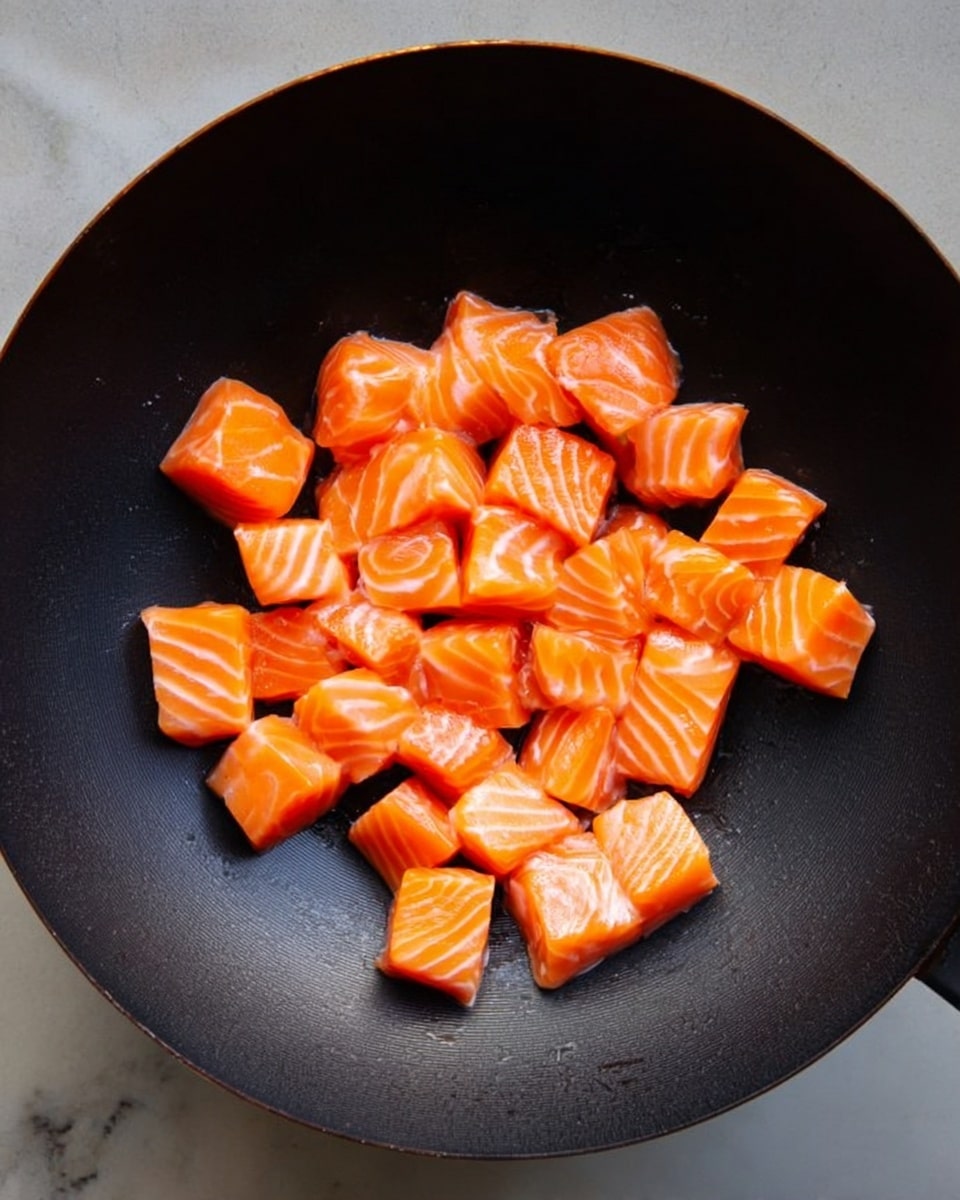 The image shows a black wok filled with evenly sized, bright orange salmon cubes. Each cube has light white lines of fat running through it, giving a fresh and rich texture. The salmon pieces are piled loosely in the center of the wok, creating a small mound with some space around the edges. The wok is set against a white marbled surface, adding a clean and simple background. photo taken with an iphone --ar 4:5 --v 7