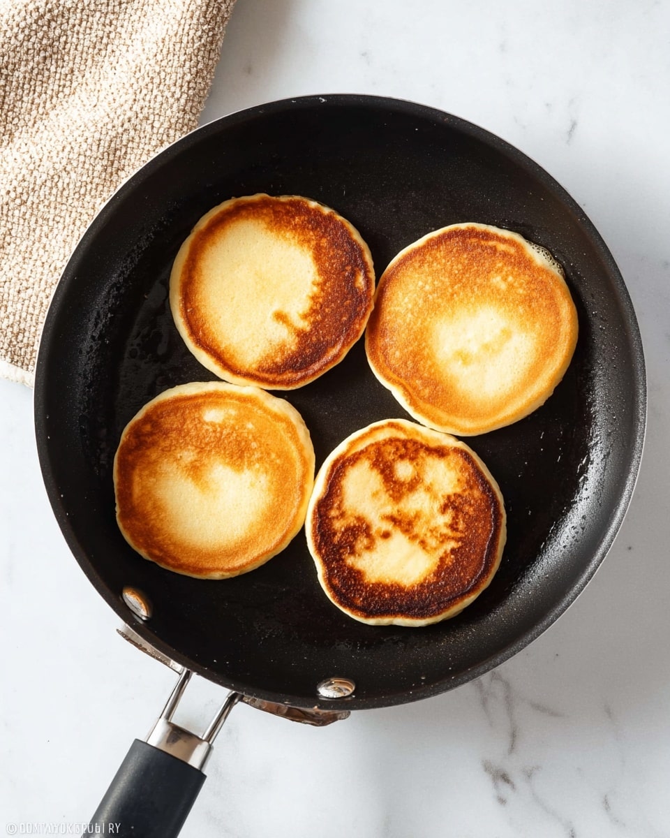 A black frying pan with a dark, smooth cooking surface contains four round pancakes, each with a golden-brown top that shows slight variations in hue and texture. The pancakes are evenly spaced in a loose square pattern inside the pan. The pan has a black handle with a slight shine, positioned diagonally to the top right. The pan sits on a white marbled surface, and a small area of a beige textured cloth is visible in the top left corner. Photo taken with an iphone --ar 4:5 --v 7