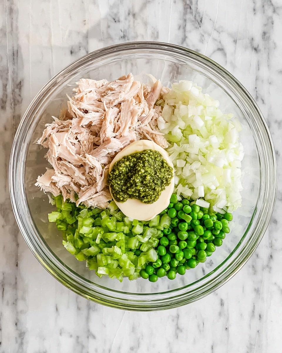 A clear glass bowl sits on a white marbled surface, filled with five main layers of ingredients arranged side by side: pale pink shredded cooked chicken on the left, next to bright green chopped celery at the top, finely chopped white onion on the right, and bright green peas at the bottom right. In the middle top of the bowl, there is a smooth pale cream layer of mayonnaise topped with a textured bright green dollop of pesto sauce. Photo taken with an iphone --ar 4:5 --v 7