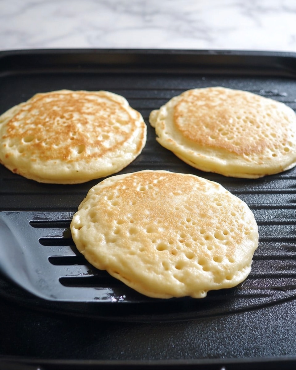 Three round, fluffy pancakes cooking on a black grill surface with light golden bubbles forming across their tops. The pancakes are pale yellow with a soft, slightly bumpy texture, and the one on the left is being lifted with a black spatula. The background shows a white marbled texture beneath the grill. photo taken with an iphone --ar 4:5 --v 7