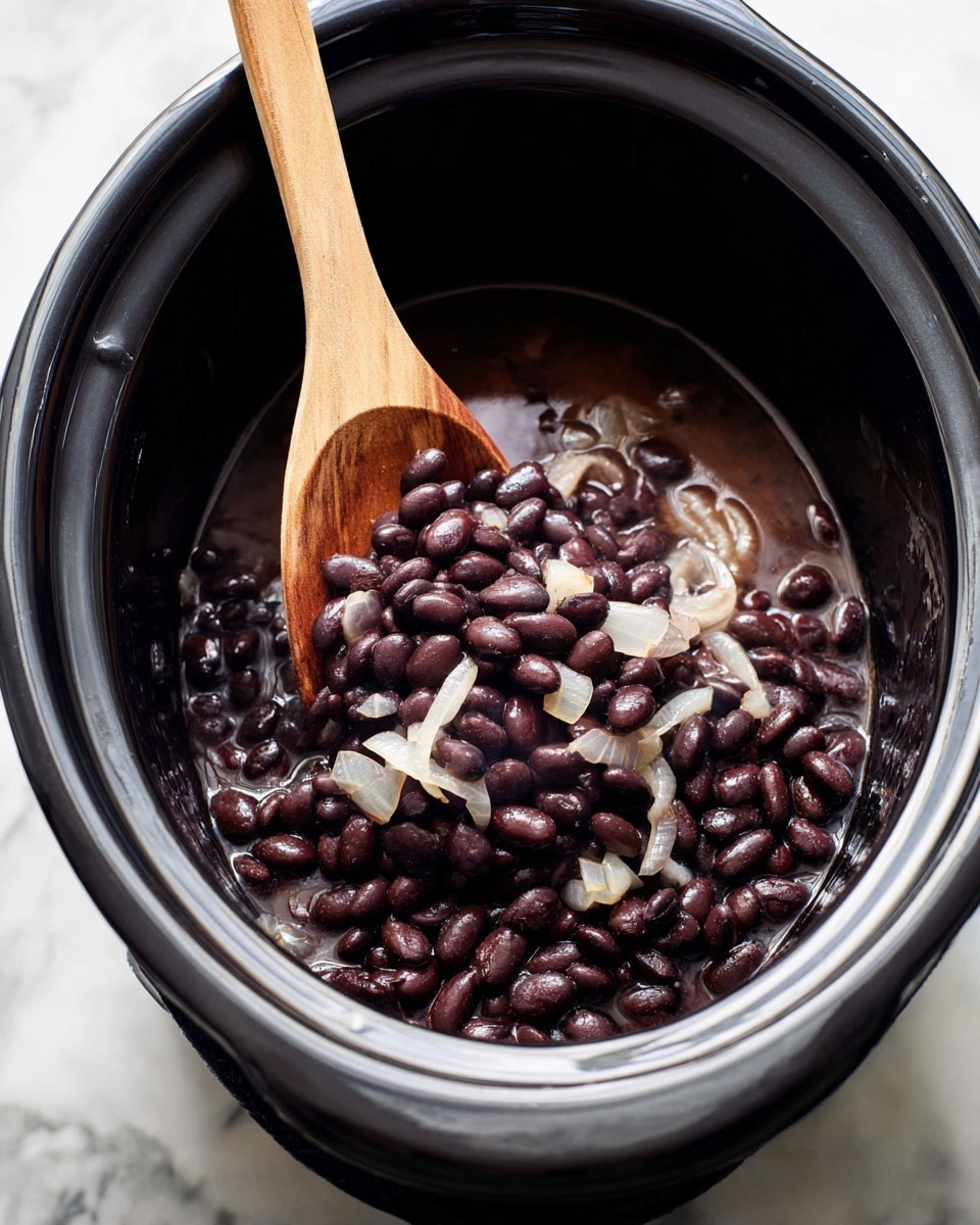 Inside a black slow cooker, there is one main layer of dark purple-black cooked beans soaking in a thin dark brown liquid. Mixed in this layer are translucent cooked onion pieces showing soft white and light brown colors with a slightly shiny texture. A light wood spoon with a smooth texture is partially dipped into the beans, resting on top of them. The slow cooker is placed on a white marbled surface. Photo taken with an iphone --ar 4:5 --v 7