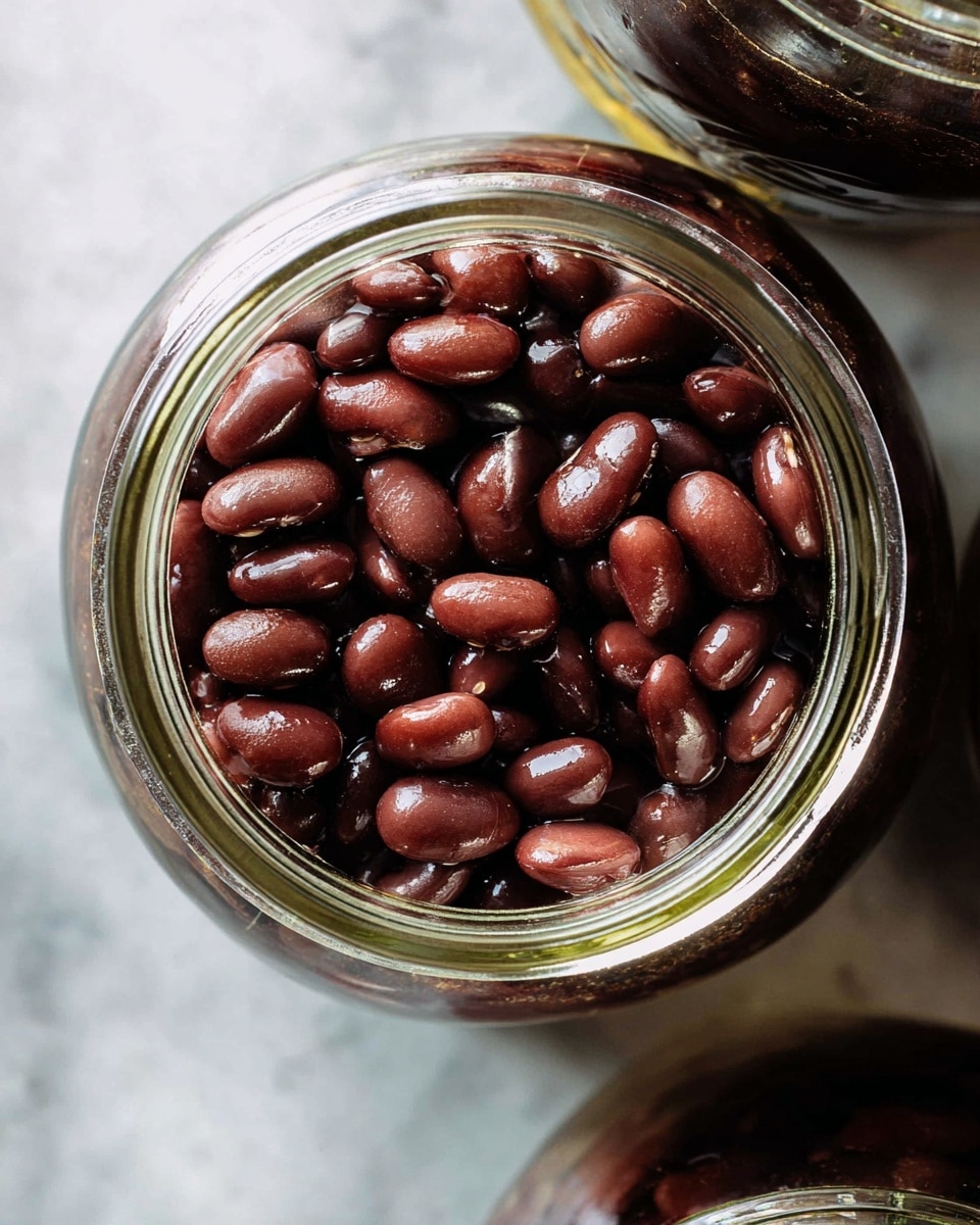 A close-up top view of a glass jar full of dark reddish-brown cooked beans filling all inside the jar visible from the top, showing a shiny wet texture and smooth round edges on the beans. The jar is placed on a white marbled surface, with part of another jar visible at the top edge. photo taken with an iphone --ar 4:5 --v 7