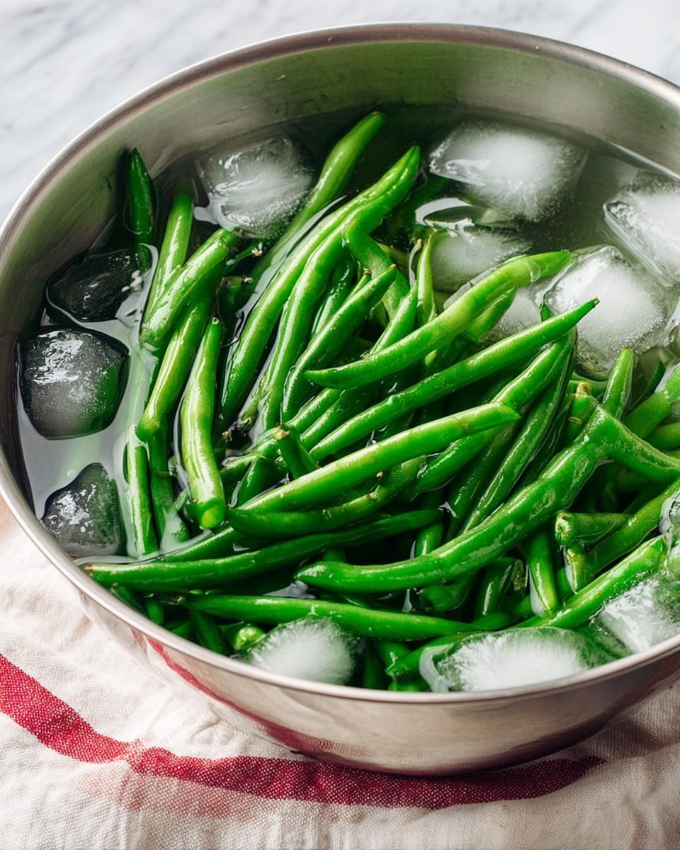 A shiny metal bowl holds fresh green beans fully covered with clear water and mixed with many ice cubes, inside the bowl, the green beans are bright and smooth, showing different angles and sizes with some curved and some straight, the ice cubes add white and transparent rough textures around the beans, the bowl is placed on a light-colored cloth with a red stripe on a white marbled surface photo taken with an iphone --ar 4:5 --v 7