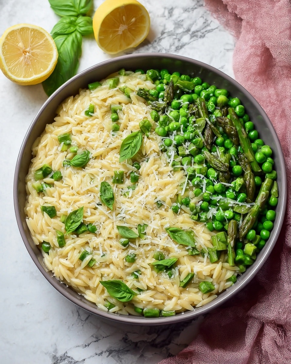 A close-up view of a white bowl filled with three layers of food: the base layer is creamy light yellow orzo pasta, the middle layer consists of bright green peas and chopped green asparagus pieces, and the top layer is scattered green basil leaves and finely grated white cheese. Next to the bowl, two lemon halves rest on a white marbled surface with a soft pink cloth in the background. Photo taken with an iphone --ar 4:5 --v 7