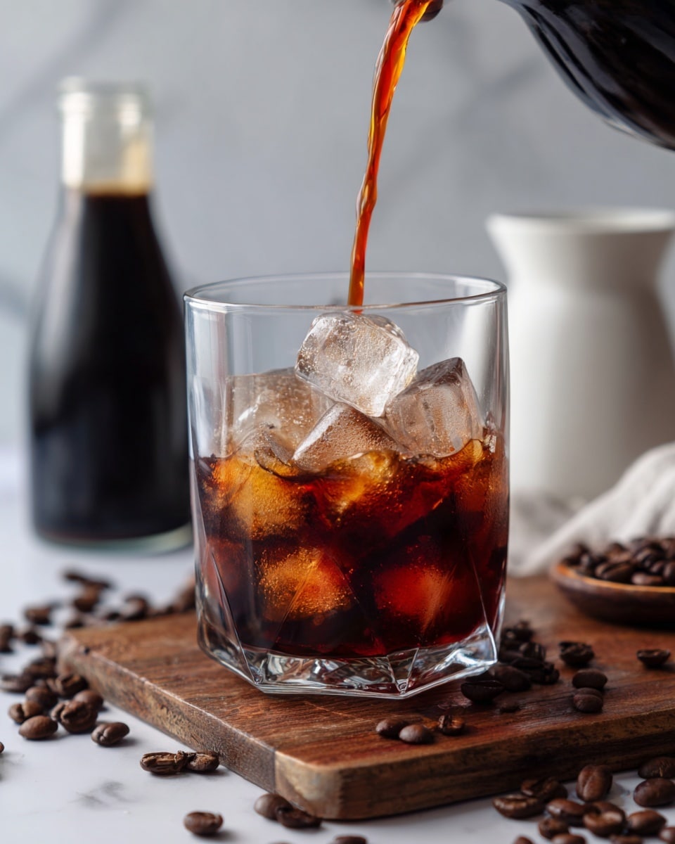 A clear glass filled with large, shiny ice cubes is being poured with dark brown cold coffee, forming a rich amber swirl as it meets the ice. The glass sits on a wooden board, surrounded by scattered dark coffee beans. In the blurred background, there is a dark glass bottle and a white ceramic container on a white marbled surface, creating a clean, bright setting. photo taken with an iphone --ar 4:5 --v 7