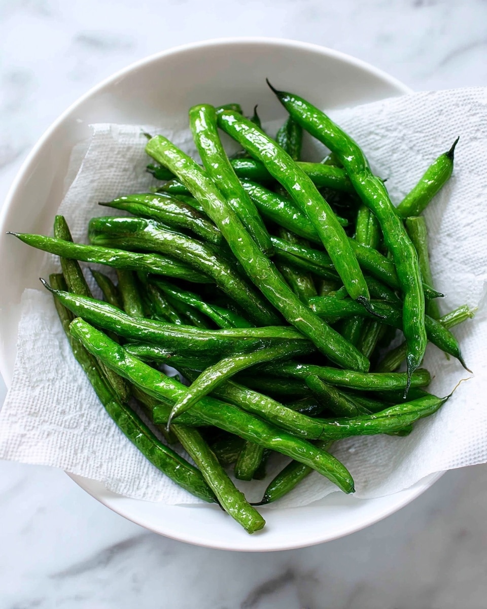 A white bowl holds a pile of cooked green beans that are bright green and slightly shiny, showing a wrinkled texture. The green beans are placed on a white paper towel inside the bowl, which absorbs extra moisture. The beans are arranged in different directions, filling the center of the bowl. The background is a white marbled surface. photo taken with an iphone --ar 4:5 --v 7