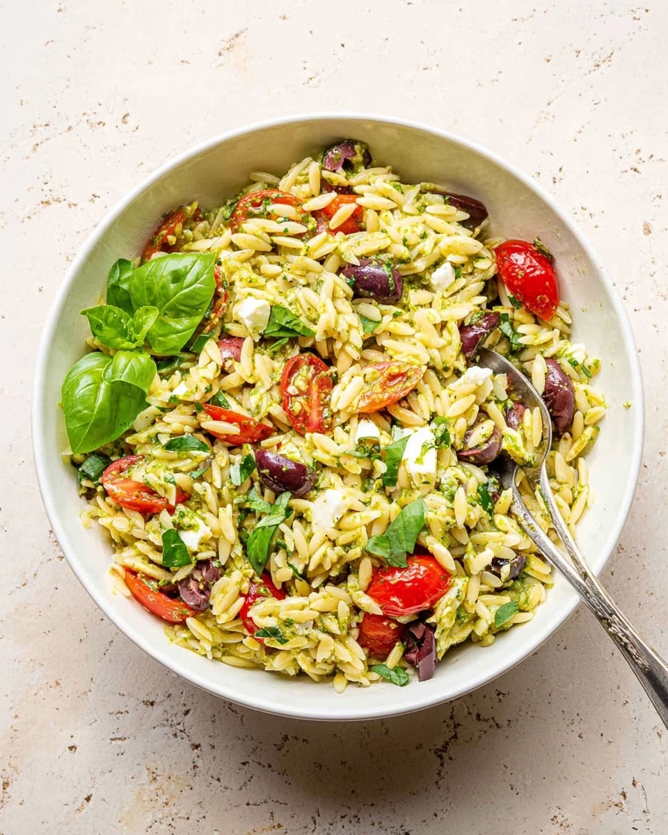 A white bowl filled with a colorful orzo salad sits on a white marbled surface. The salad has multiple layers with small yellow orzo pasta forming the base mixed with diced green herbs and pesto, scattered bright red cherry tomato halves, dark purple olive slices, and small white pieces of cheese. On top, fresh green basil leaves are spread unevenly. A silver spoon rests inside the bowl, leaning towards the left. The image is bright with natural light, showing the texture of the pasta and the freshness of the ingredients photo taken with an iphone --ar 4:5 --v 7