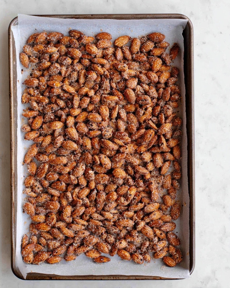 The image shows a baking tray lined with white parchment paper, filled with a single layer of roasted almonds. The almonds are coated in a light, slightly grainy mixture that looks like it contains spices and sugar, giving a slightly rough texture with small dark specks visible across the surface. The almonds are golden-brown with some darker toasted spots, and they are spread evenly to cover the entire tray. The baking tray sits on a white marbled textured surface, and the photo gives a clear, close-up view from above. photo taken with an iphone --ar 4:5 --v 7