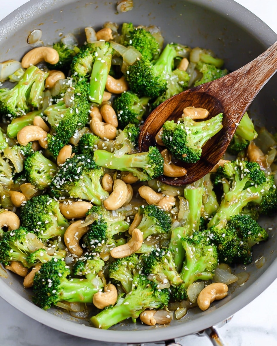 A close-up view of a light gray frying pan filled with a stir-fry of bright green broccoli florets, light golden cashew nuts, and translucent cooked onion slices. A wooden spoon is resting in the pan, holding some broccoli and cashews. White sesame seeds are scattered over the top, adding small white details. The pan is set on a white marbled surface. photo taken with an iphone --ar 4:5 --v 7
