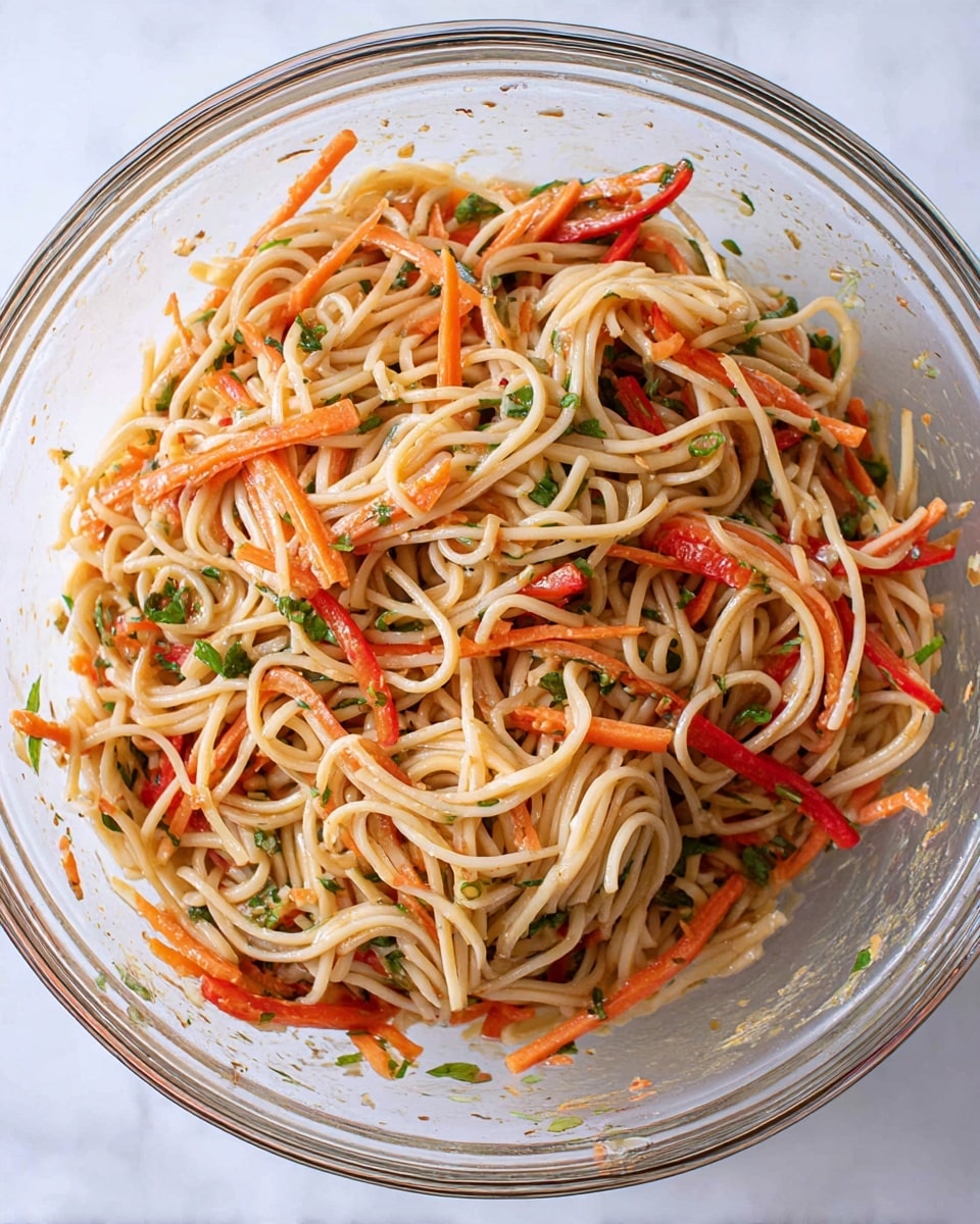 The image shows a clear glass bowl filled with mixed noodles and julienned vegetables on a white marbled surface. The base layer is pale beige noodles with a smooth texture, intertwined with thin, bright orange carrot sticks and red bell pepper strips scattered evenly throughout. Small green herbs and chopped scallions add spots of green color, mixed well among the noodles and vegetables. The mixture has a light coating of sauce that gives a slight sheen to the ingredients. The bowl is cleanly presented with some residue of sauce on the glass sides. Photo taken with an iphone --ar 4:5 --v 7
