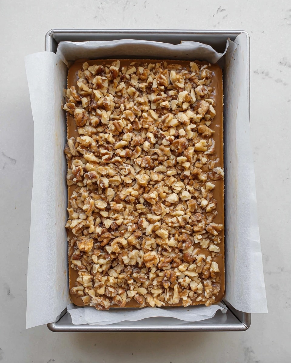 A rectangular cake with one main layer of light brown batter, topped generously with a thick layer of chopped walnuts in various sizes, creating a rough, textured surface. The cake is in a metal baking tray lined with white parchment paper, with the paper edges slightly crinkled and folded outward. The tray sits on a white marbled surface, and the photo is taken from above, showing the whole cake and tray clearly. photo taken with an iphone --ar 4:5 --v 7