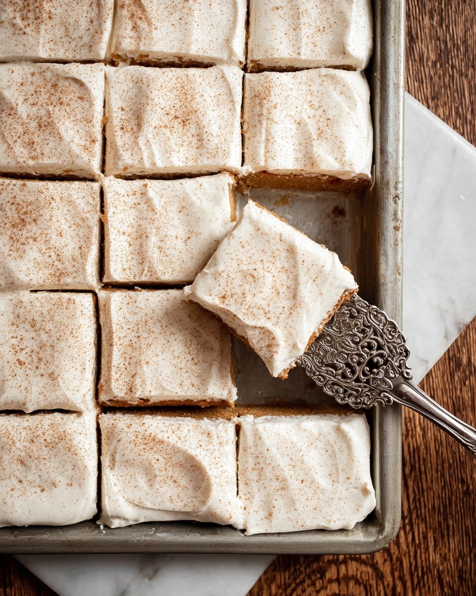 A rectangular metal baking tray holds a sheet of cake cut into twelve square pieces with light brown cake base as the first layer, topped with a thick, creamy white frosting spread smoothly and evenly. Each frosted square is sprinkled with light brown powder on top. One piece is lifted on a silver ornate spatula on the right side, showing the soft texture of the cake and creamy layer above, and the tray is set on a white marbled surface. Photo taken with an iphone --ar 4:5 --v 7