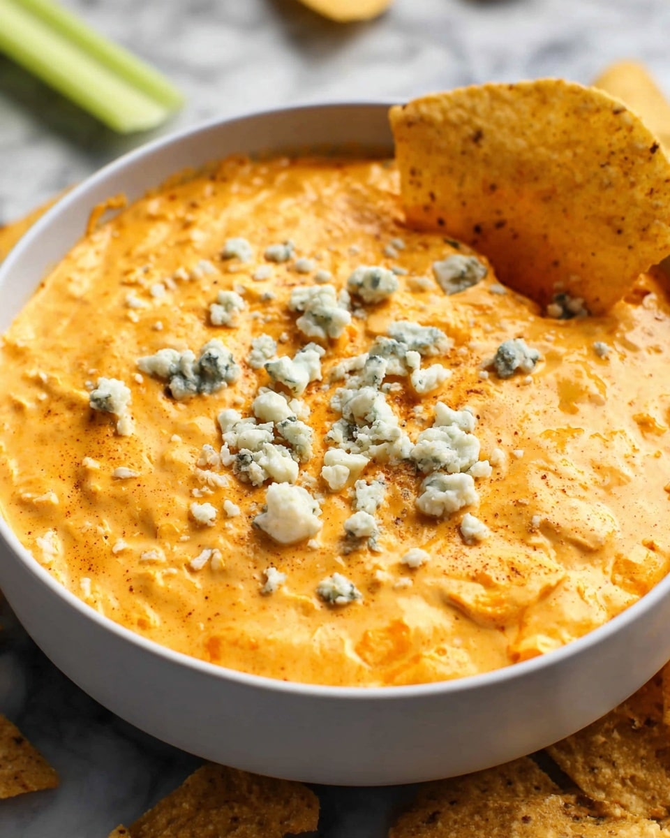 A white bowl filled with thick, creamy orange cheese dip with a slightly rough texture, topped with small, irregular crumbles of blue cheese scattered on the surface; a crispy tortilla chip is dipped into the right edge of the bowl. The bowl sits on a white marbled surface with some blurred celery sticks and more tortilla chips around the edges. photo taken with an iphone --ar 4:5 --v 7