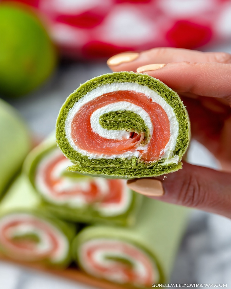 The image shows a close-up of a rolled wrap held by a woman's hands with light peach nail polish. The wrap has three visible layers: the outer and inner layers are green with a soft, sponge-like texture, and between them is a thick white creamy layer, followed by a smooth pinkish-red layer resembling smoked salmon, rolled tightly to create a spiral pattern. In the background, multiple similar wraps are stacked, slightly blurred, on a white marbled surface, with a green round object and a red and white checkered pattern also blurred in the far background. Photo taken with an iphone --ar 4:5 --v 7