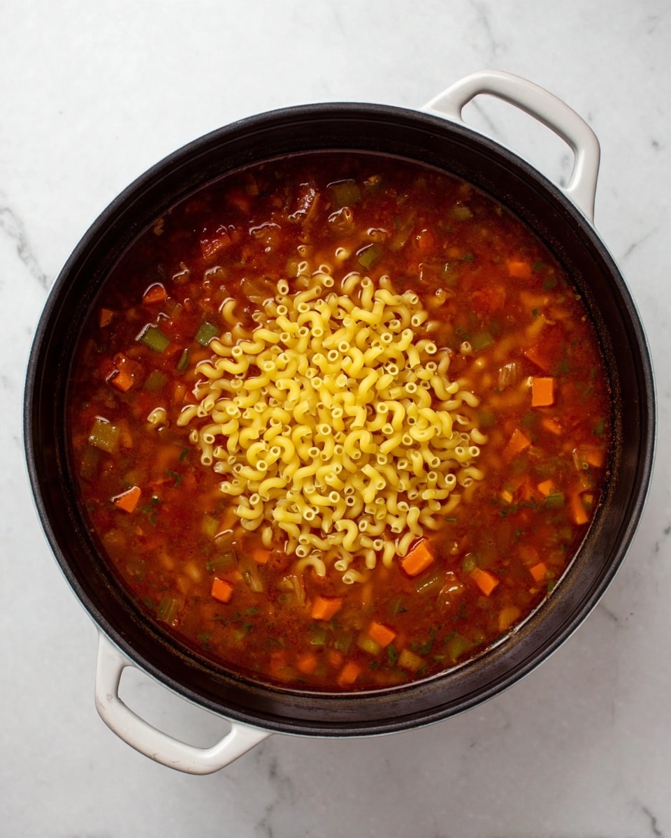 A black pot with white handles is filled with a reddish soup base that has small pieces of vegetables like orange carrots and green bits floating in it. In the middle of the pot, there is a pile of dry yellow elbow macaroni pasta placed on top of the soup, standing out against the darker liquid. The pot is set on a white marbled surface, giving a clean and simple background. photo taken with an iphone --ar 4:5 --v 7