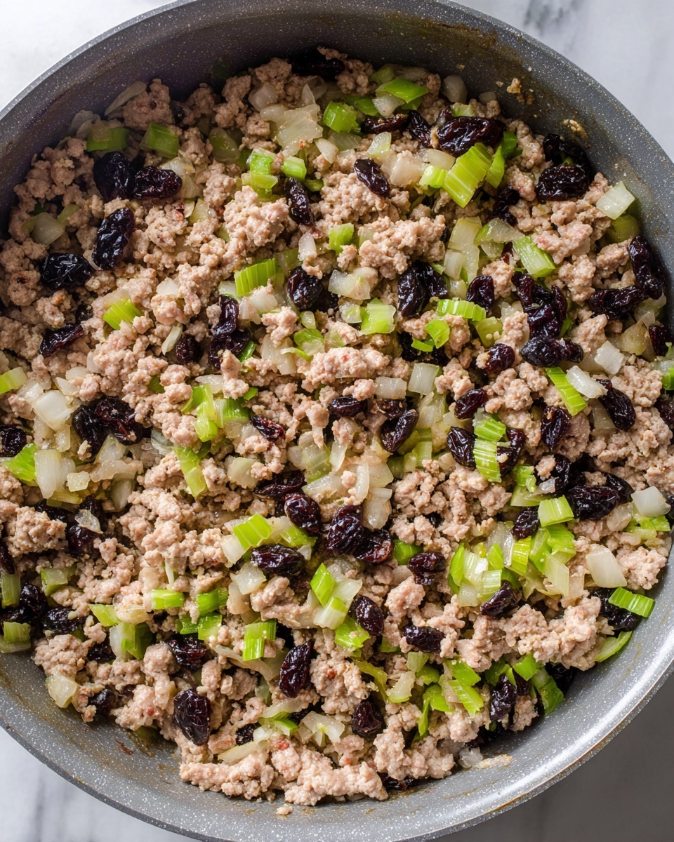 The image shows a close-up of cooked ground meat mixed with small diced celery pieces, chopped onions, and dark dried berries or raisins. The meat is light brown and crumbly, spread evenly with green celery bits and translucent white onion pieces scattered throughout. The dark dried fruit adds contrast with their shiny deep purple-black color. This mixture is inside a gray pan on a white marbled surface. The textures include soft meat, crunchy celery, and chewy dried fruit, all closely packed inside the pan. photo taken with an iphone --ar 4:5 --v 7