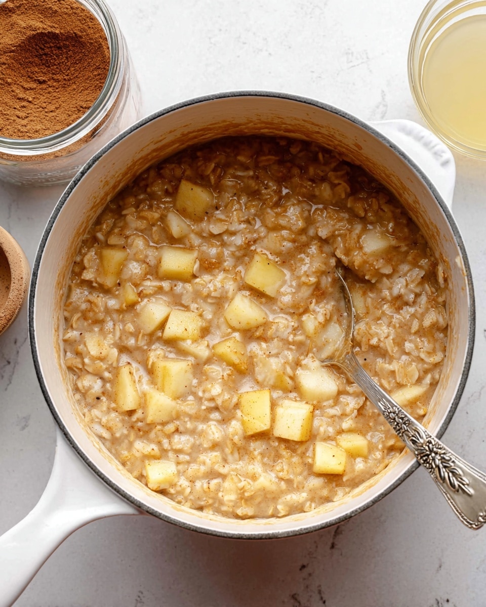 A white pot filled with a thick oatmeal mixture that has a light brown color with visible chunks of soft, pale yellow apple pieces mixed throughout. The texture looks creamy with small bits of oats clearly seen. A silver spoon with a decorative handle is partially submerged on the right side of the pot. The pot sits on a white marbled surface, next to a glass jar filled with cinnamon powder and a glass bowl with a pale yellow liquid. photo taken with an iphone --ar 4:5 --v 7